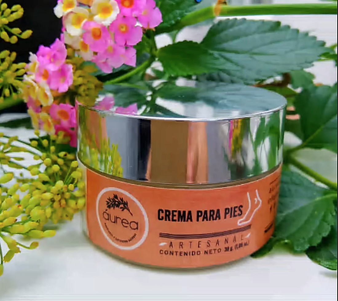 Crema para pies artesanal de Aurea Herbolaria, en un frasco de tapa plateada entre flores