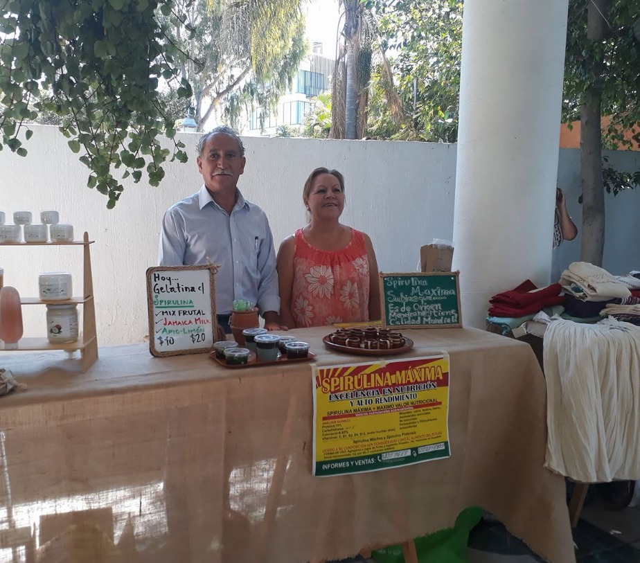 Agustín Bautista y su esposa en el puesto de Spirulina Máxima, junto a frascos del producto y un cartel con el análisis nutricional