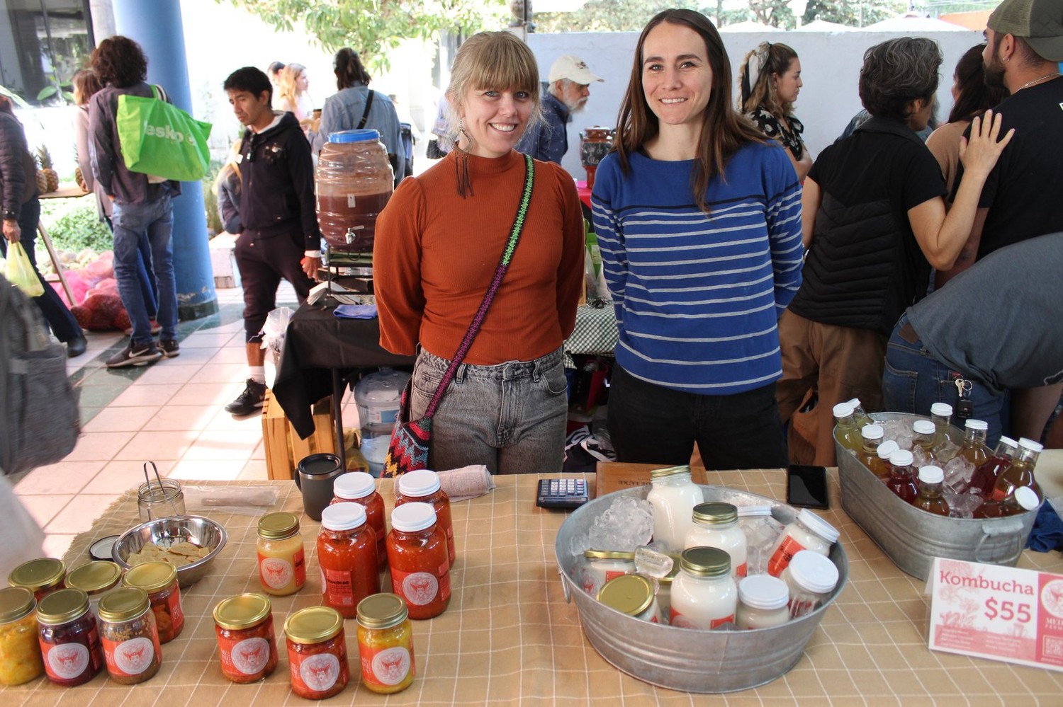 Las dos productoras de Medusa Fermenta detrás de su puesto, con un garrafón de kombucha en proceso y los frascos de fermentados en la mesa