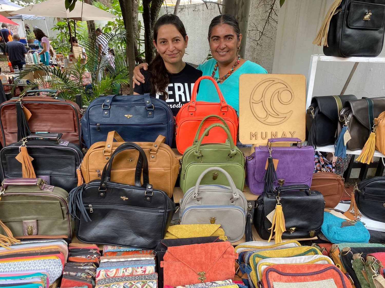 Las dos productoras de Munay detrás de su puesto, con decenas de bolsas y carteras de colores y el logo de madera tallado