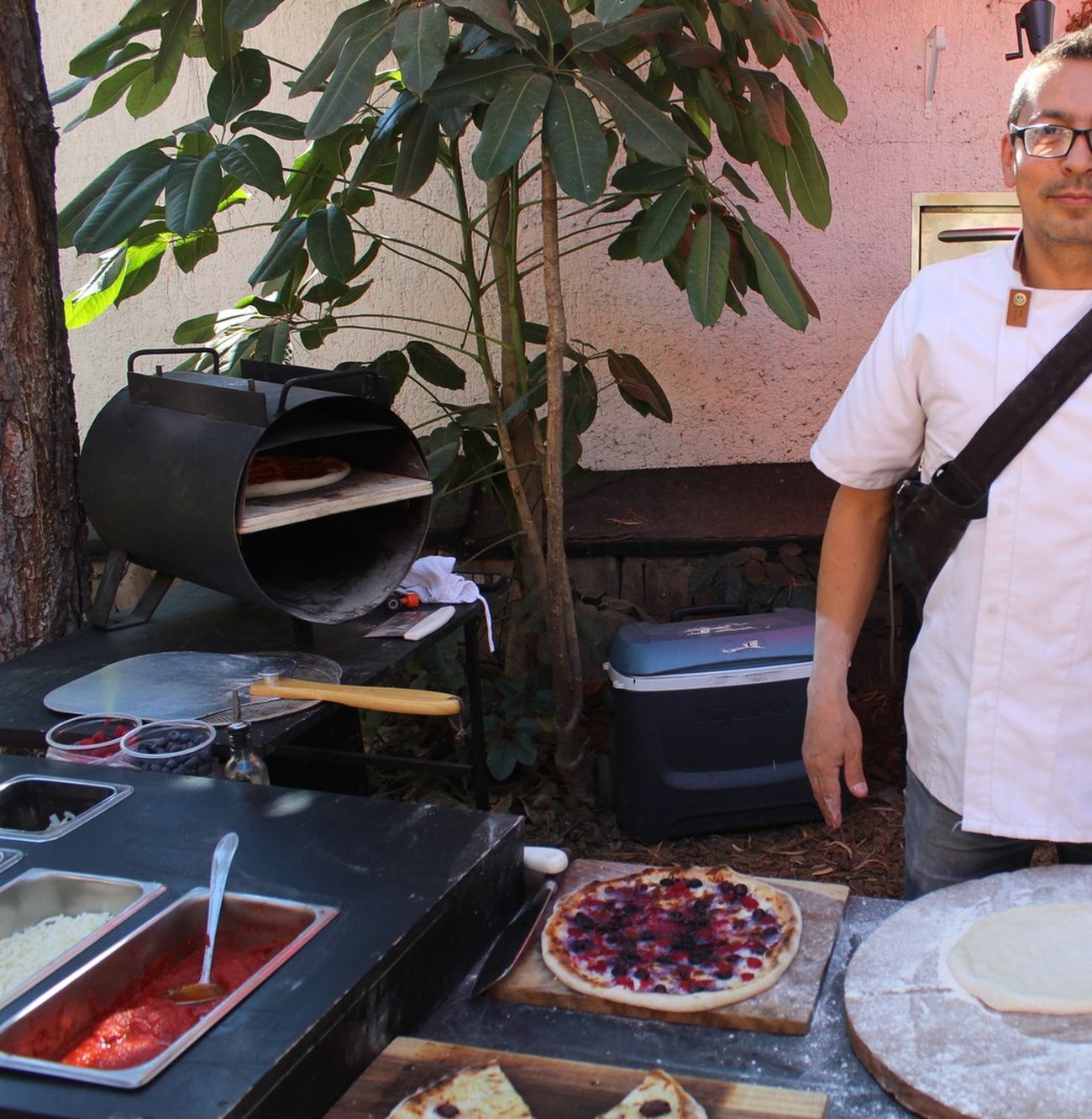 Pizzero de Pizza Di Strada junto a su horno de leña cilíndrico, con una pizza horneándose dentro