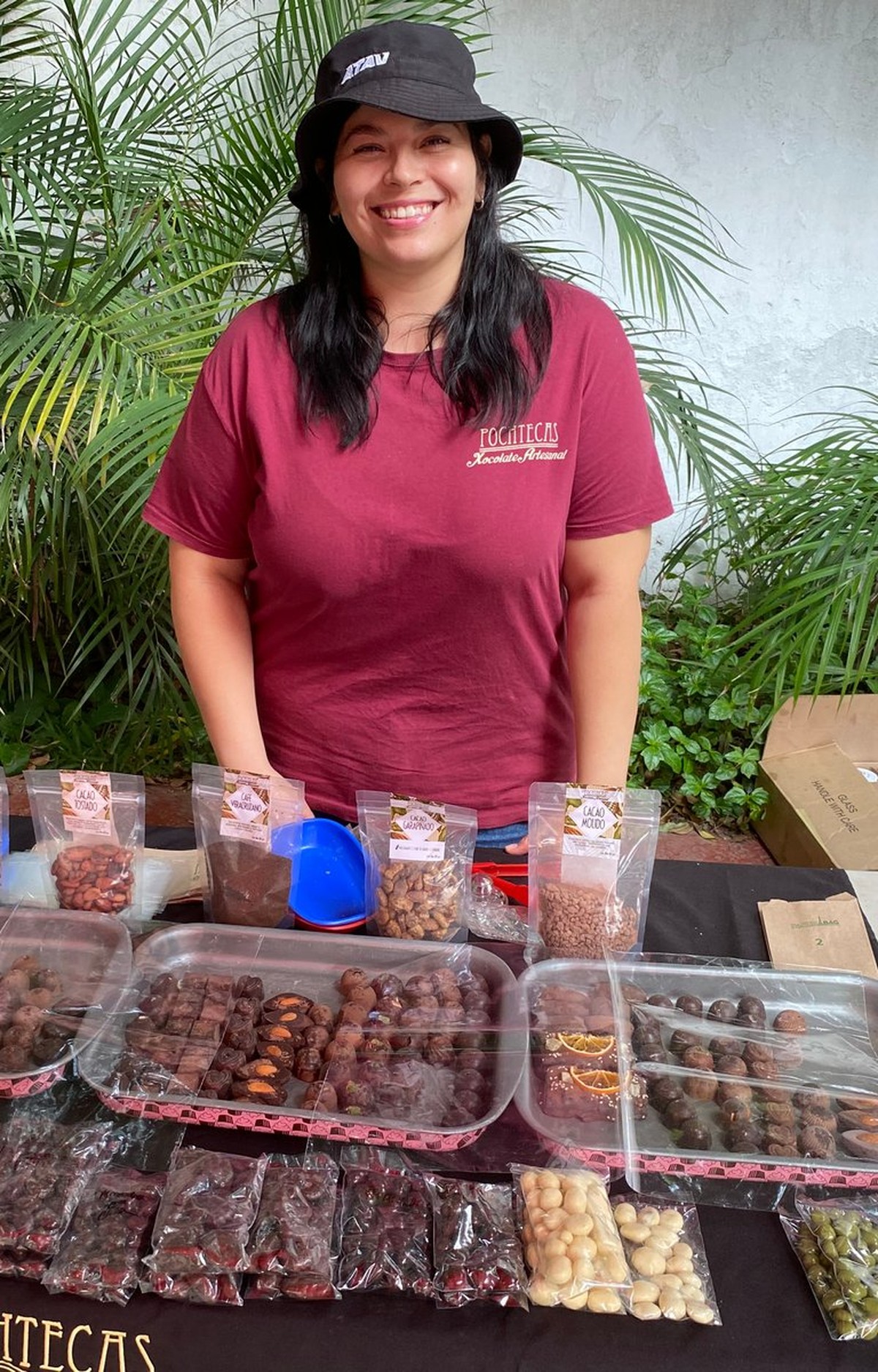 Vendedora de Pochtecas con playera de la marca, detrás de charolas de trufas y bombones de chocolate y bolsas de cacao