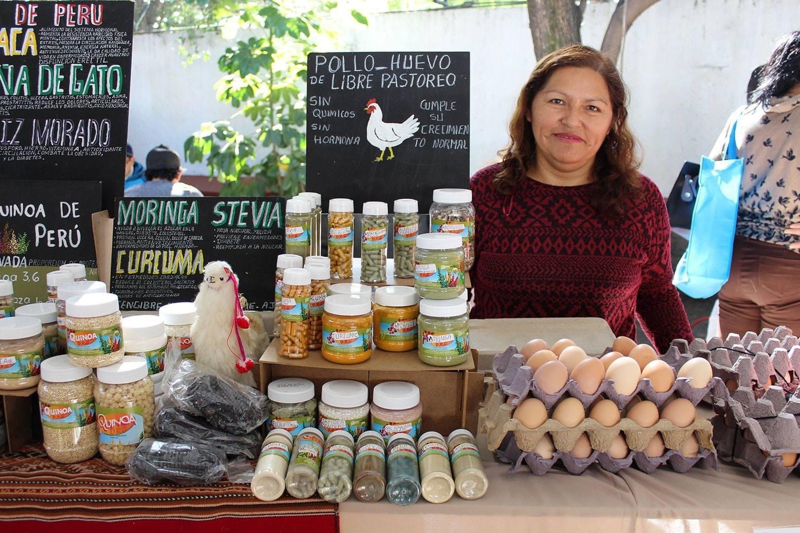 Devora Retamoza Guillén en el puesto de Productos Deby, con frascos de moringa, stevia y quinoa, y charolas de huevo