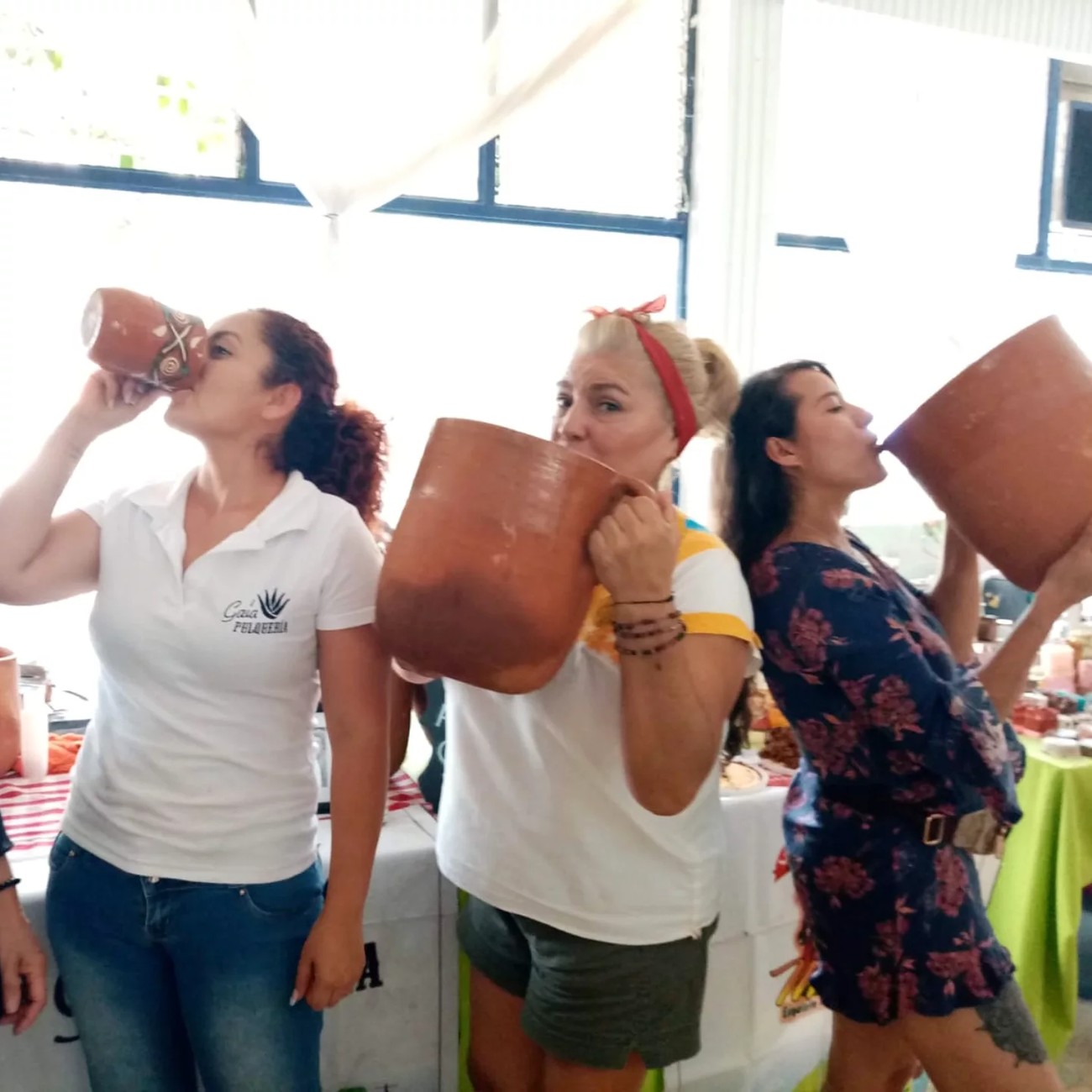 Tres mujeres bebiendo pulque de jarros de barro en una cata; la del polo bordado 'Gaia Pulquería' al frente