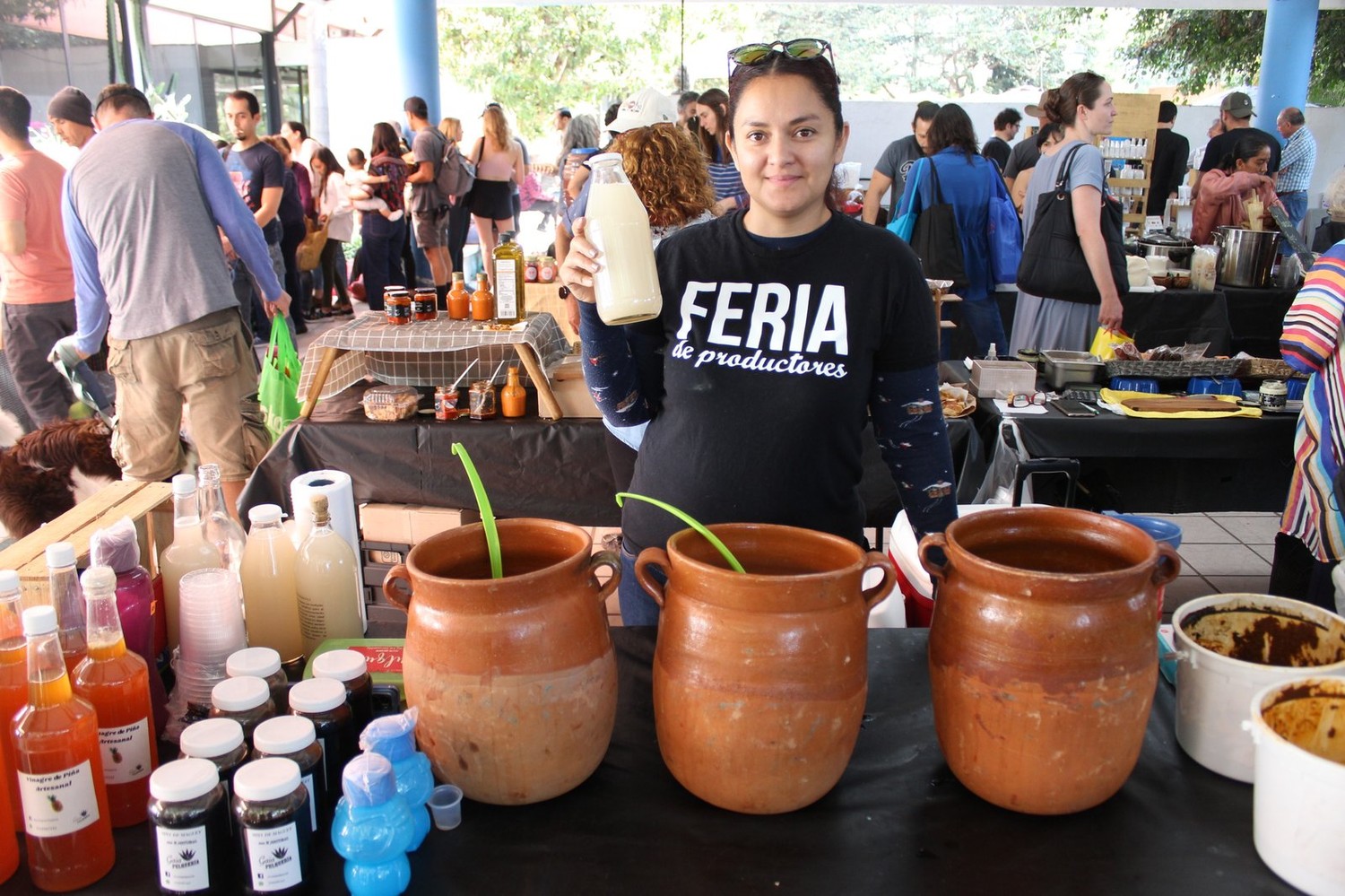 Vendedora de Pulquería Gaia con playera 'Feria de productores', sosteniendo una botella de aguamiel junto a tres ollas de barro de pulque